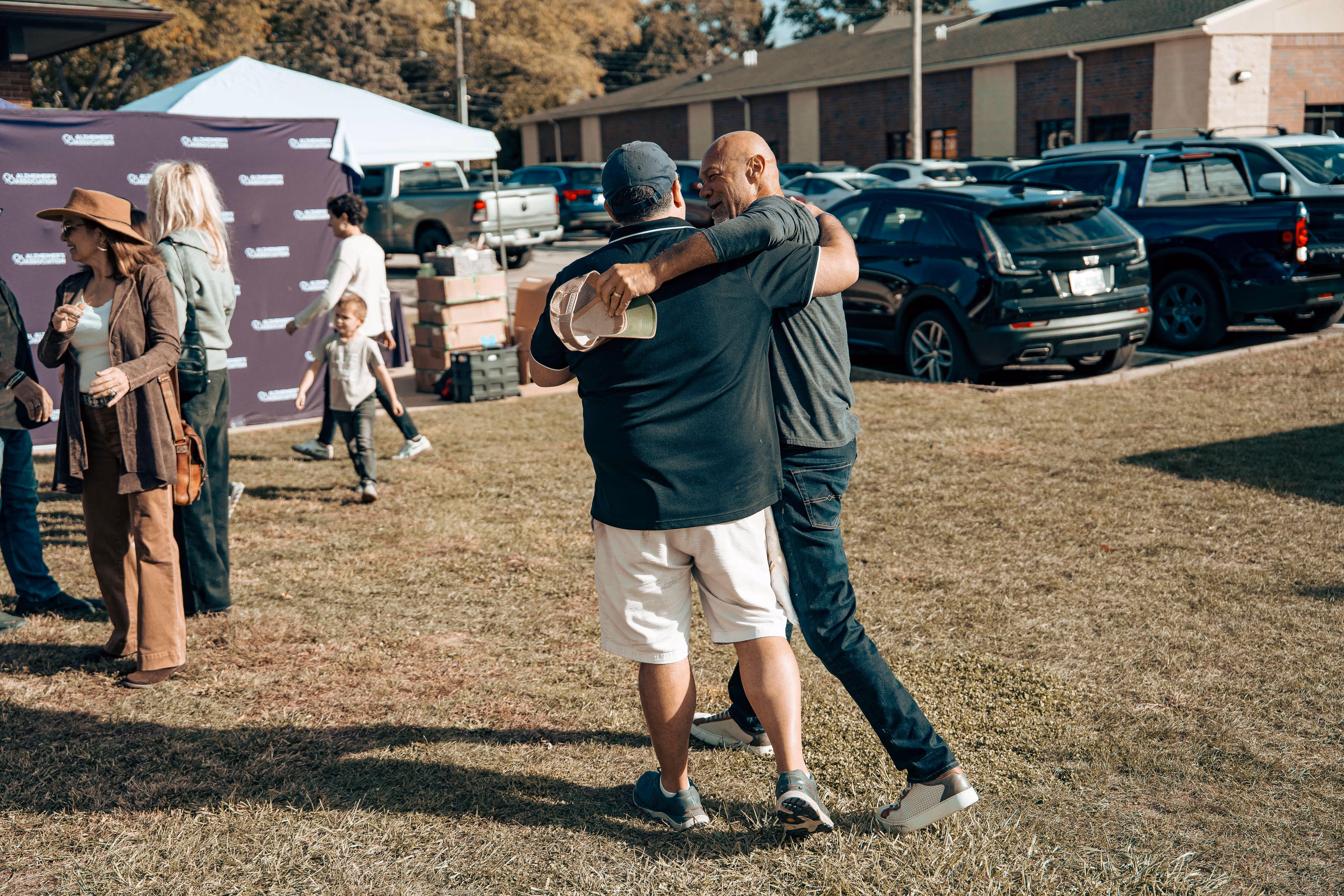 Two men hugging on a grassy area with parked cars and event tent
