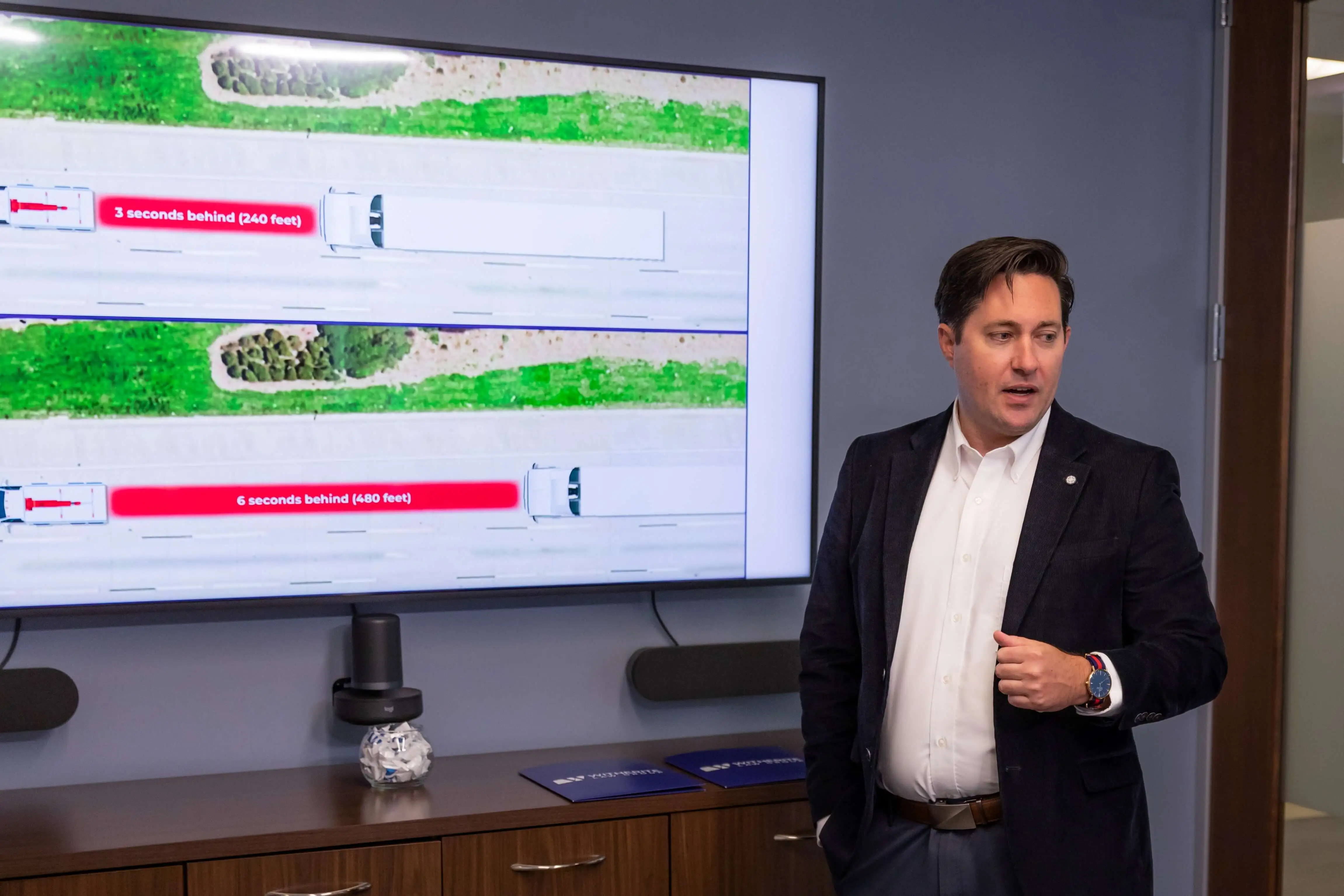 A traffic safety expert lecturing during a presentation, standing next to a large screen displaying a visual aid illustrating truck following distances and how they contribute to a truck wreck.