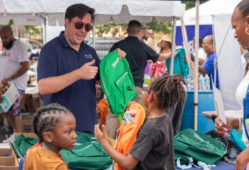 Man donating green backpacks to children at an outdoor community outreach event. The man is wearing a blue polo shirt and sunglasses, giving a thumb-up gesture while talking to the kids.
