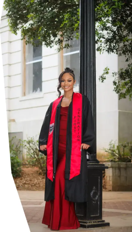 A young female graduate wearing a black gown over a long red dress. She has a red and black stole that is embroidered with "FIRST GENERATION." She is posing outdoors next to a black lamp post in front of a white building.