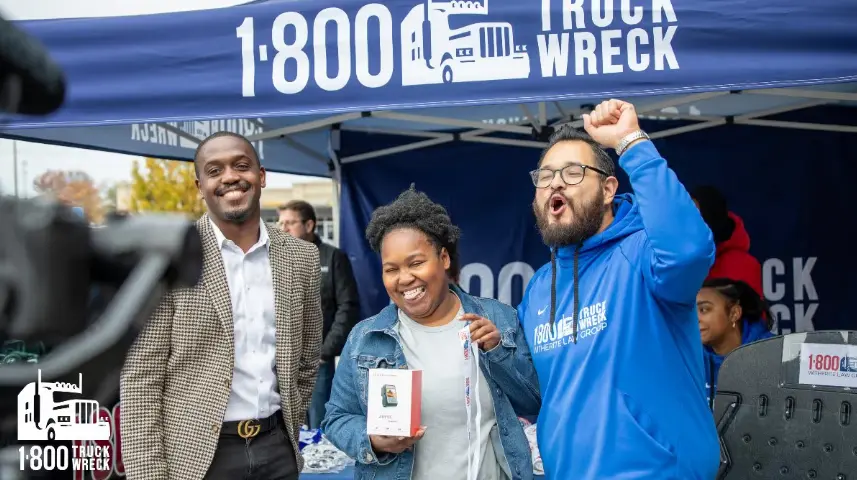 A celebratory moment at an outdoor event under a large blue tent labeled "1-800 TRUCK WRECK." Three smiling people, two men and a woman holding a small boxed prize, are cheering, with one man enthusiastically raising his fist.