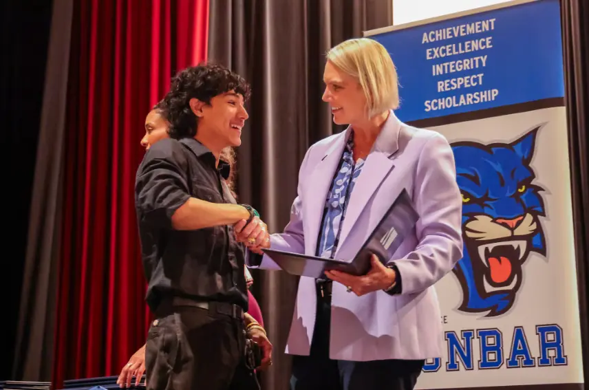 Amy Witherite, wearing a light lavender blazer, smiles and shakes hands with a young male student in a dark shirt on a stage. They are at an awards ceremony, with a banner for Dunbar featuring the text "ACHIEVEMENT EXCELLENCE INTEGRITY RESPECT SCHOLARSHIP" and a blue panther logo visible behind them.
