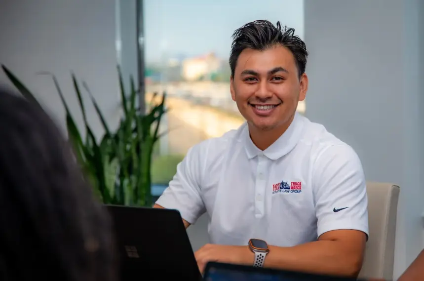 A smiling young man with dark hair, wearing a white polo shirt with a company logo, is seated in a brightly lit office setting. A blurred outdoor scene is visible through the window behind him.