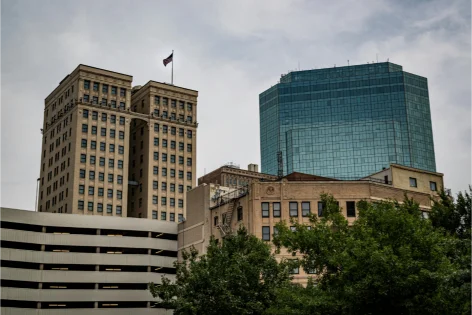 Skyline view contrasting historic stone buildings with a contemporary, blue-glass modern structure. A small American flag is visible atop the older building.