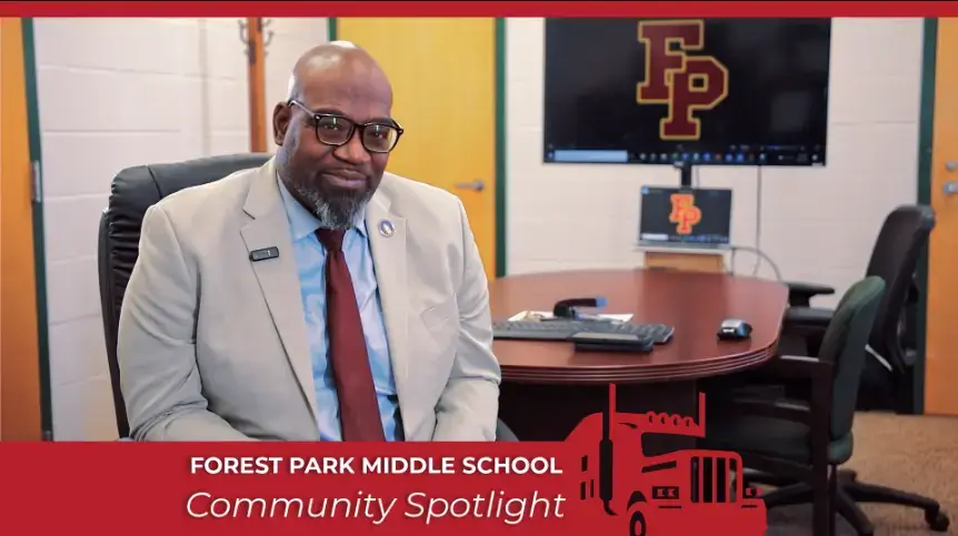 Community Spotlight interview with a man in his office at Forest Park Middle School. The segment title is displayed below, and the school logo is visible on a monitor behind him.