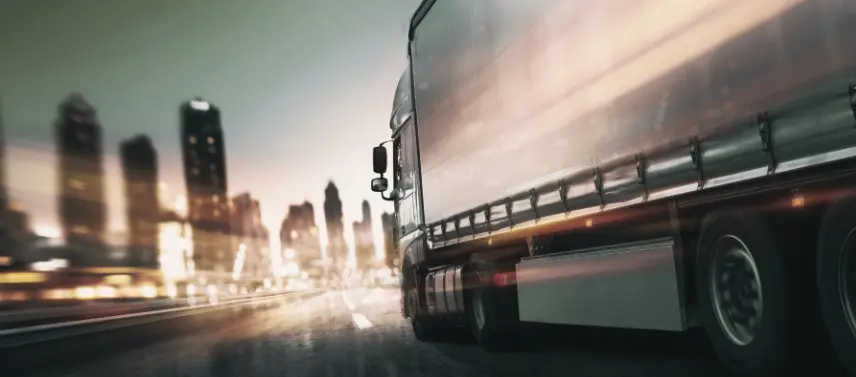 Semi-truck traveling at high speed on a road at night, captured with motion blur against a bright, stylized city skyline backdrop.