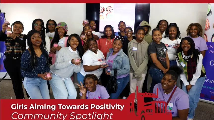 Group photo of young girls and mentors participating in a "Girls Aiming Towards Positivity" community spotlight event. The group is standing together and smiling indoors.