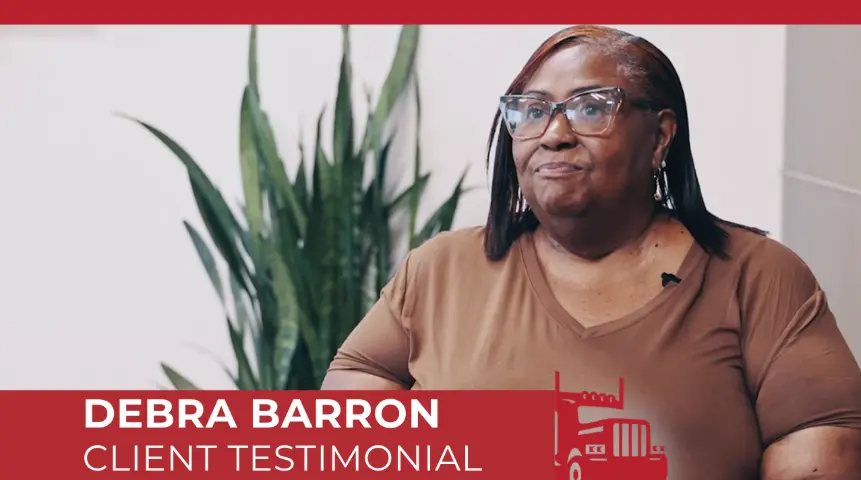 Debra Barron giving a client testimonial video interview, seated indoors and wearing glasses and a brown top, with a green houseplant visible in the background. 