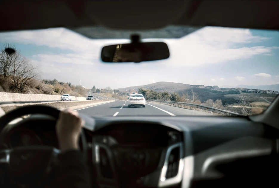 Driver's view perspective from inside a car, showing the steering wheel and open road on a highway with distant hills visible under a bright sky.