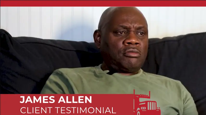 James Allen giving a client testimonial video interview with a serious expression, seated on a dark couch. 