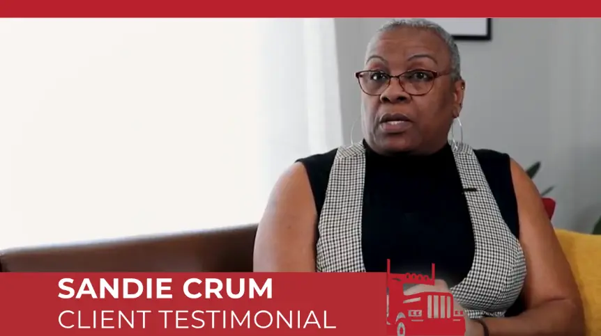 Sandie Crum giving a client testimonial video interview, wearing glasses and a plaid vest over a black top.