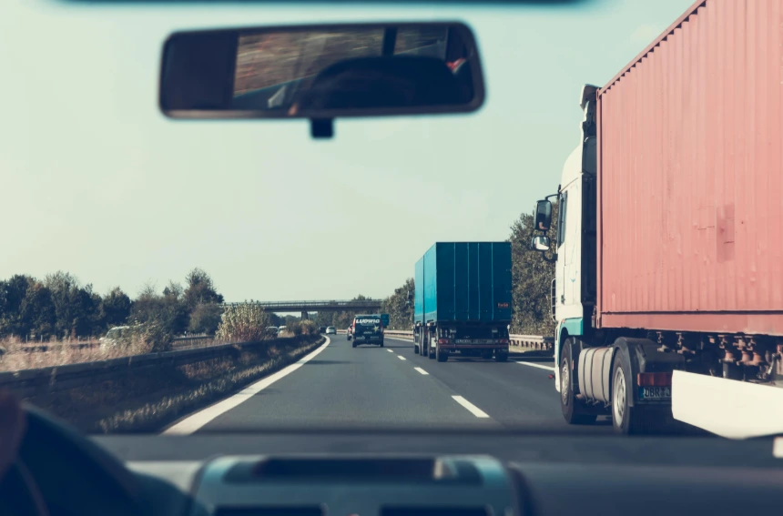 The driver's view from inside a car on a highway, showing multiple commercial trucks ahead, including a large red and blue container truck. 