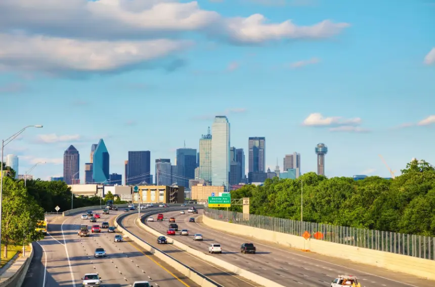 Busy highway traffic leading toward the Dallas, Texas, skyline, featuring the Reunion Tower and downtown skyscrapers under a blue sky.