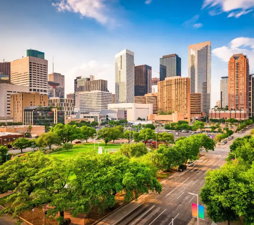 Panoramic view of the vibrant Houston, Texas, downtown skyline rising above a lush green park area and tree-lined streets on a sunny day.