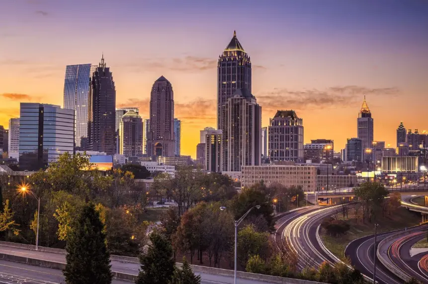 The downtown Atlanta, Georgia skyline at sunset, featuring prominent skyscrapers illuminated against an orange and purple sky. A busy highway interchange with time-lapse light trails is visible in the foreground, with trees separating the road from the bui