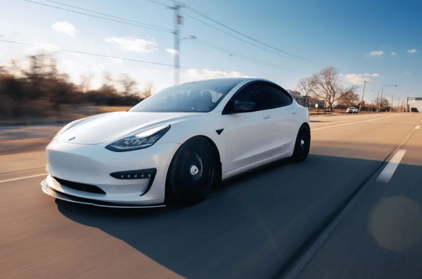 A dynamic, low-angle action shot of a white Tesla Model 3 sedan driving at speed on a multi-lane road on a sunny day. The car has dark wheels and a subtle body kit, with motion blur visible on the road surface.
