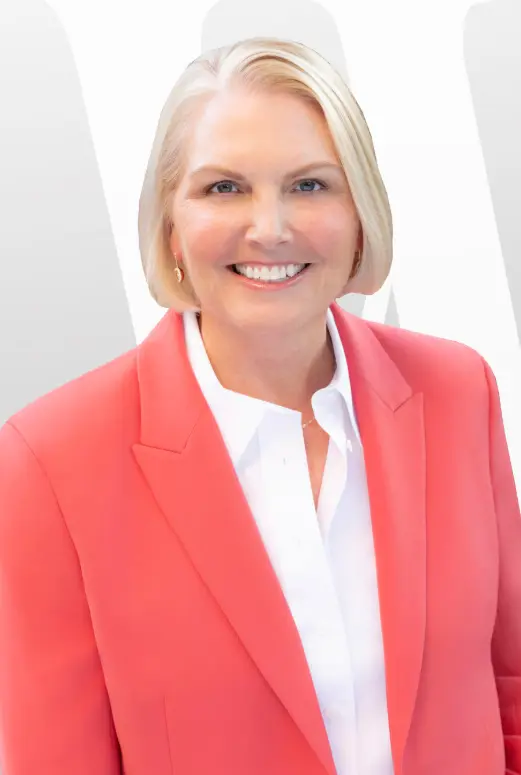 A headshot portrait of Amy Witherite, wearing a bright coral-pink blazer over a crisp white collared shirt. She is positioned against a background of white with a faint, stylized gray 'W' graphic.