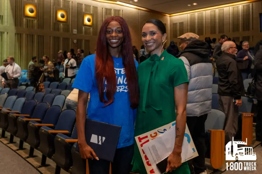 Two smiling women stand side-by-side in a large auditorium filled with blue seating. The woman on the left has long red hair and wears a blue t-shirt. The woman on the right wears a bright green dress. Both are holding folders. The 1-800-TRUCK-WRECK logo i