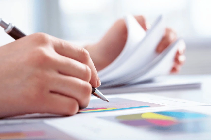 A close-up shot of a person's hand holding a pen and writing on a document that features colored pie charts or graphs, while their other hand is flipping through a stack of papers in the background.