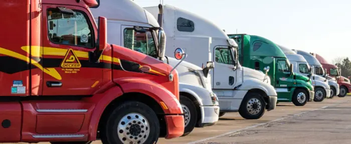 A line of commercial semi-truck cabs of various colors—including a red Decker truck in the foreground, white, and green—are parked side-by-side in an outdoor lot on a sunny day.
