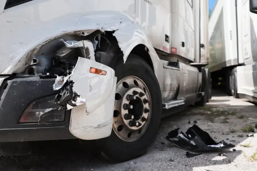 A close-up, low-angle shot of the front wheel and lower side of a damaged white semi-truck. The front fender is severely cracked and broken, with debris on the ground. Another white trailer is visible on the right.