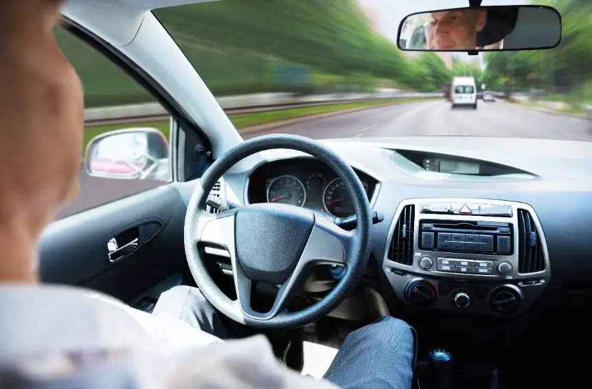 A driver's point-of-view shot from the interior of a car, focusing on the steering wheel and dashboard. The road ahead is motion-blurred, suggesting high speed. The driver's face is partially visible in the rearview mirror.