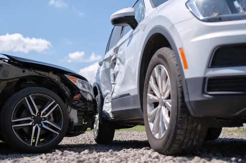 A close-up, low-angle shot of a car accident showing two vehicles that have collided. A white SUV on the right has severe damage to the driver's side door panel. A dark or black car on the left has major front-end damage with its bumper missing.