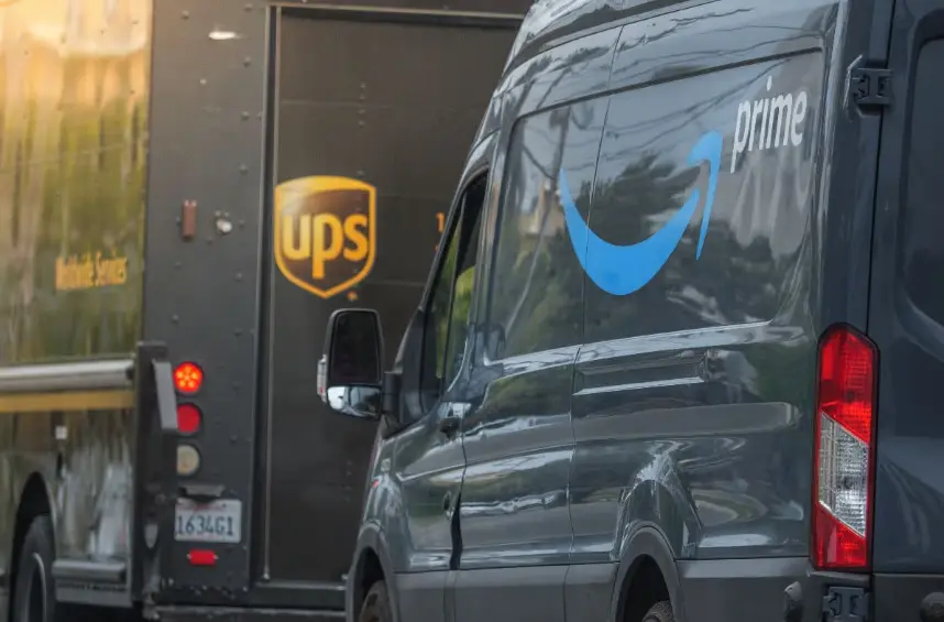 A close-up of a dark gray Amazon Prime delivery van with the blue "prime" logo on its side, passing or parked next to the back of a brown UPS delivery truck. The UPS logo and license plate are visible on the truck in the background.