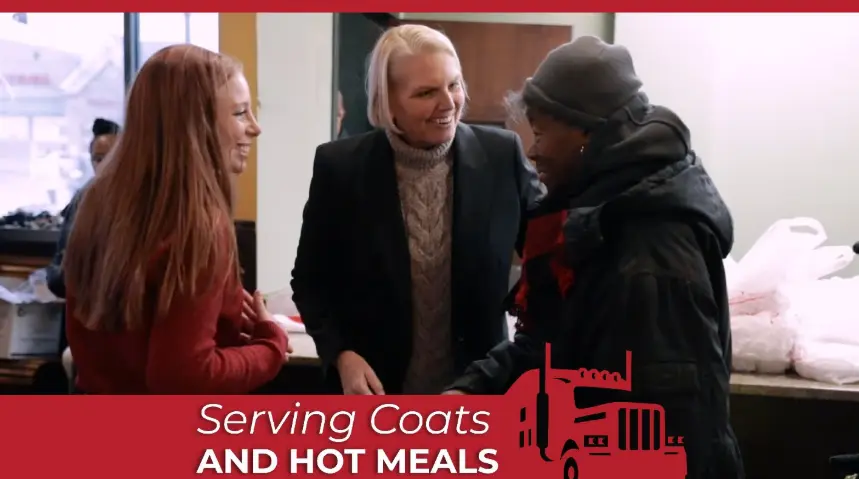 Amy Witherite, wearing a black blazer and a cowl-neck sweater, smiles while interacting with a person in a dark coat at a donation event. Another woman with long red hair smiles on the left. A red banner across the bottom reads: "Serving Coats and Hot Meal