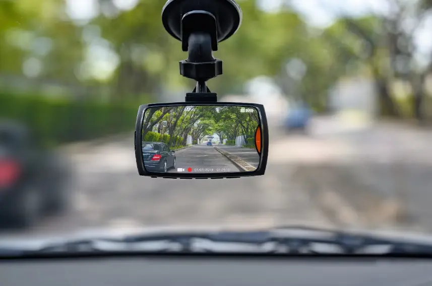 A close-up view of a car dashcam mounted on the windshield, clearly showing its screen recording a tree-lined street ahead. The image demonstrates using a dashcam for accident evidence.
