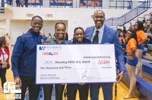 Four smiling people—two adults and two younger individuals—hold a large novelty check for "$21,000" made out to the "Munday Mills H.S. Band" for the Great Atlanta Band Challenge. The check features the Witherite Law Group and 1-800-TRUCK-WRECK logos, prese