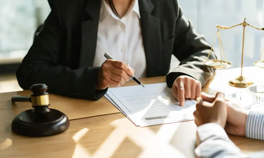 A close-up shot of a lawyer (wearing a dark suit) pointing with a pen to a section of a document while consulting with a client. A gavel and a small scale of justice are visible on the dark wooden table.
