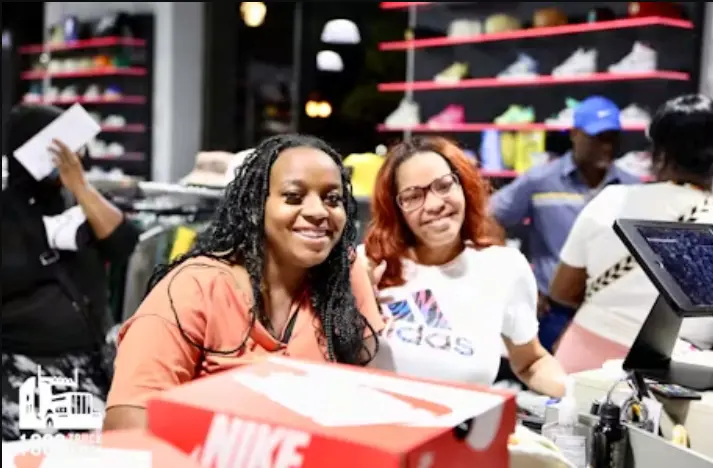 Two smiling women, one with long braids and one with red hair and glasses wearing an Adidas t-shirt, are standing behind a red Nike shoe box at a retail store counter. Shelves of sneakers are visible in the background.