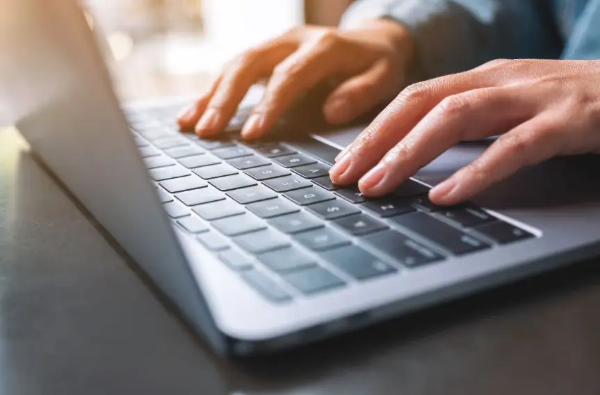 A close-up, angled shot of hands typing on the keyboard of a silver laptop in a well-lit setting, suggesting work or casual browsing.