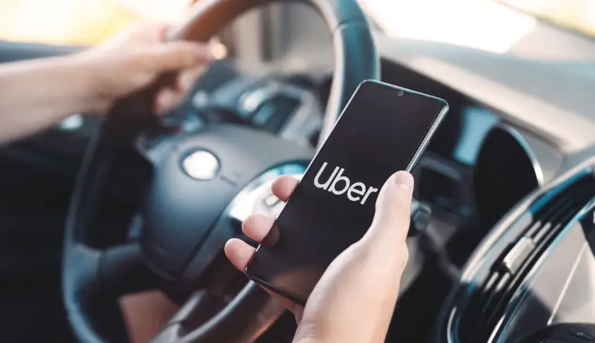 A person's hands holding a smartphone displaying the Uber app logo, while the other hand rests on the car's steering wheel. This is a driver's perspective inside a vehicle.
