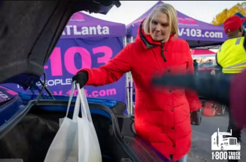 Amy Witherite, wearing a bright red puffer jacket and black gloves, places white shopping bags into the open trunk of a blue car. She is at an outdoor charity event with purple and pink tents, and the 1-800-TRUCK-WRECK logo is in the corner.