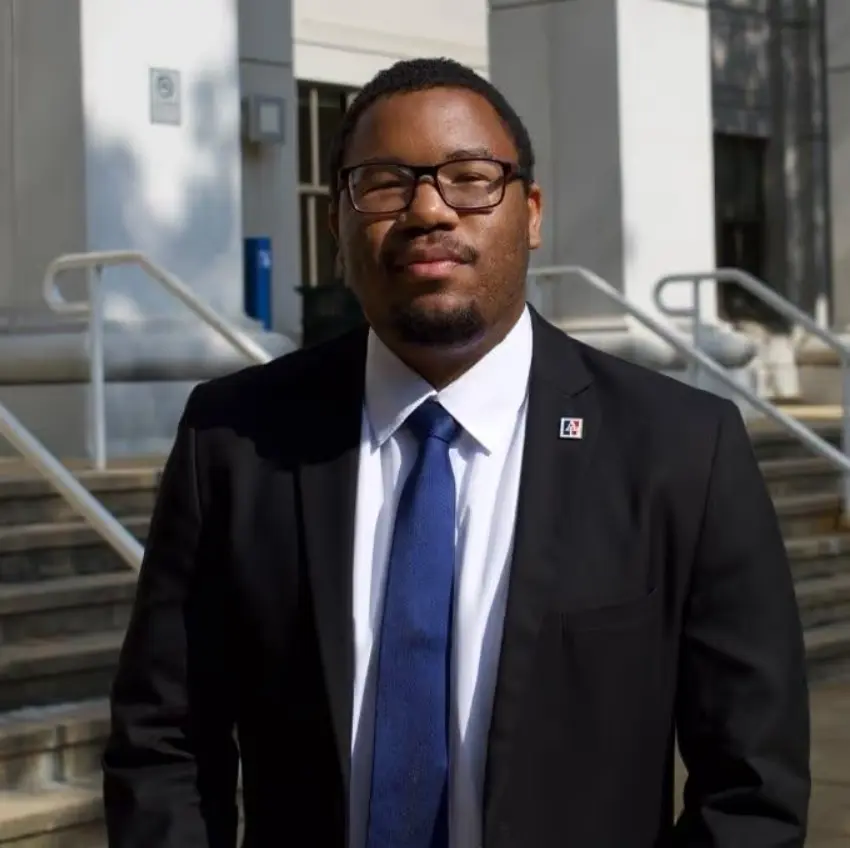 A portrait of a man wearing a black suit, white shirt, and blue tie, standing outdoors in front of the steps of a government or court building. He is wearing glasses and looking directly at the camera.