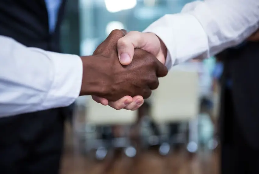 A close-up shot of two people shaking hands, one with dark skin and one with light skin, both wearing white professional shirts and dark suits. The handshake is central in the frame, with a blurred office background.