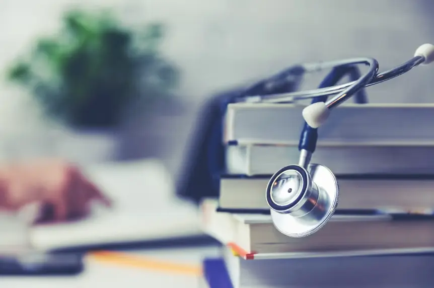 A close-up shot of a classic silver and blue stethoscope resting on a stack of medical textbooks. In the blurred background, a person is seen studying or working at a desk, emphasizing learning and medicine.
