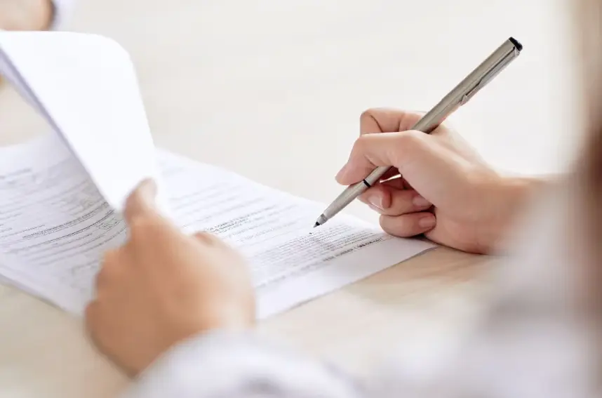 An over-the-shoulder view of a person's hand using a silver pen to sign or fill out a form or legal document. The person's other hand is holding and turning a page.