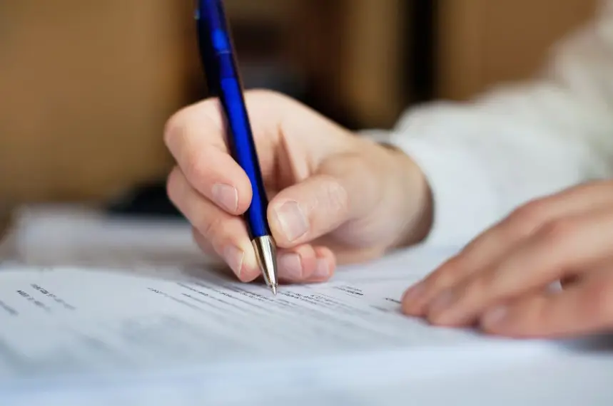 A close-up shot of a hand holding a dark blue pen and writing or signing a document on a white paper surface. The person is wearing a white cuff shirt.