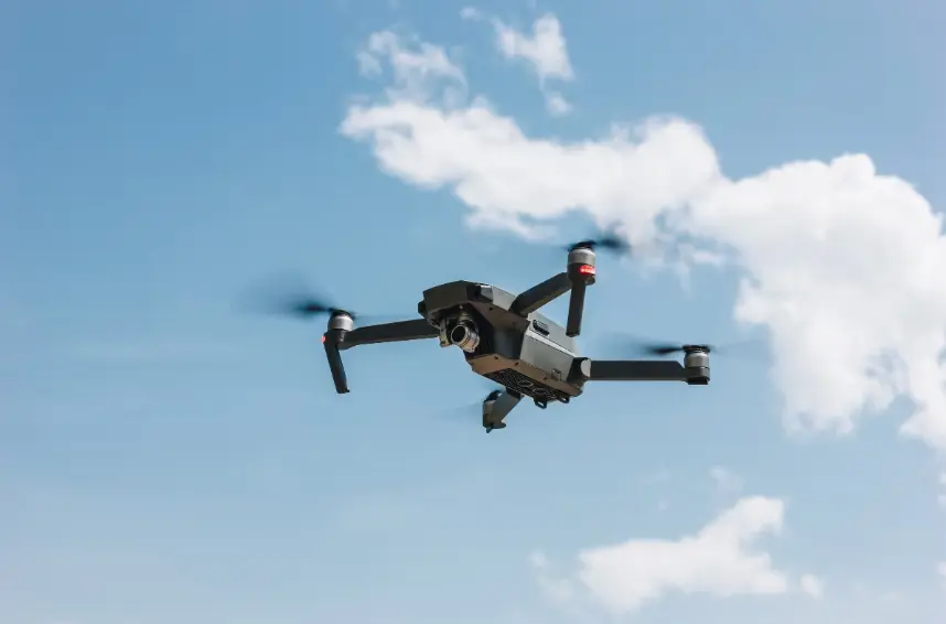 A dark gray quadcopter drone, with its camera visible beneath the body, is flying and hovering in the air against a bright blue sky with white, fluffy clouds.