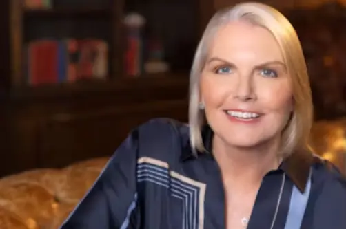 A headshot of Amy Witherite, wearing a dark navy blue silk blouse with light geometric stripes. She is seated in a dimly lit, traditional setting, possibly on a tufted leather sofa with a bookshelf in the blurred background.