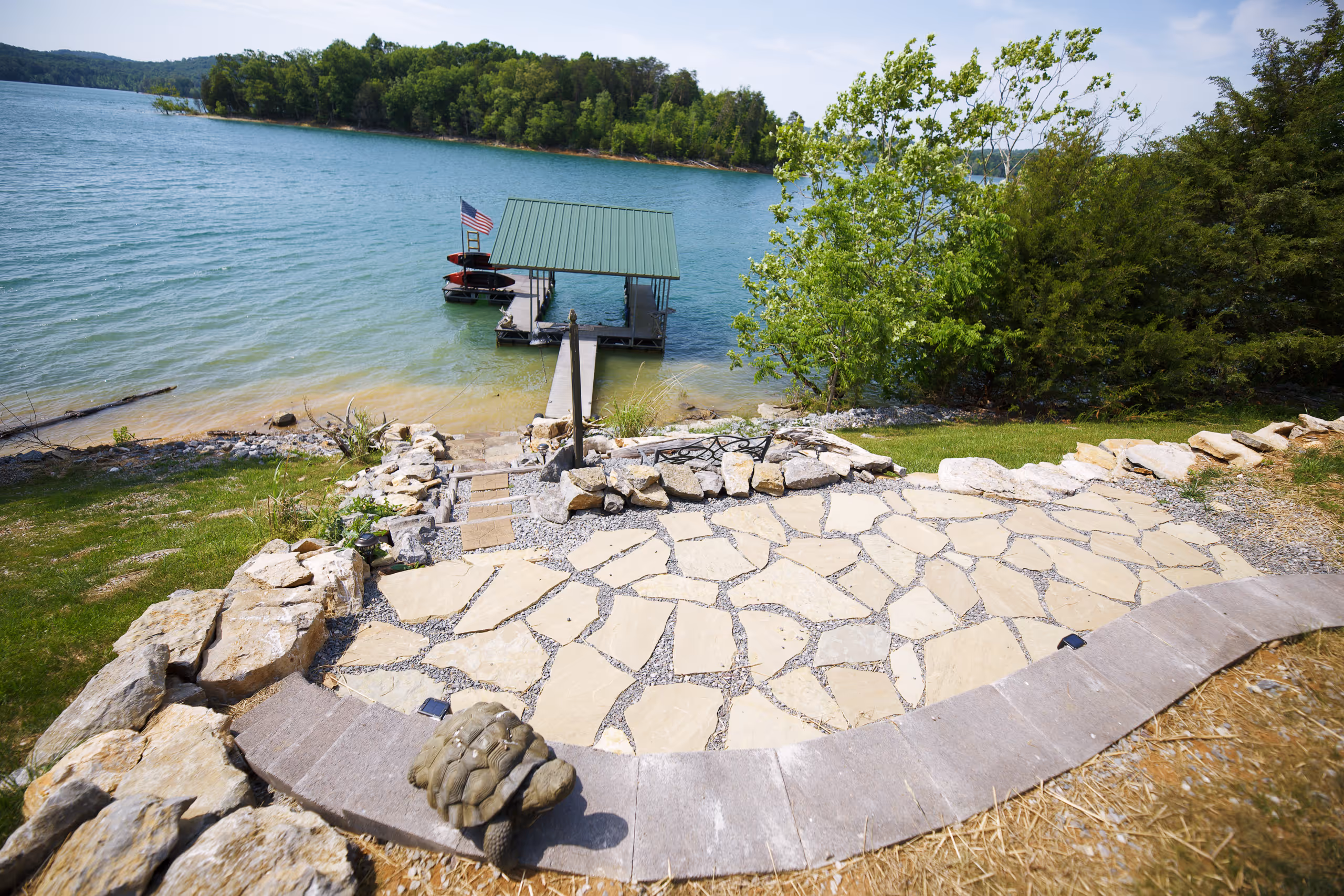 A lakeside patio with a stone walkway, a dock, and a clear blue sky