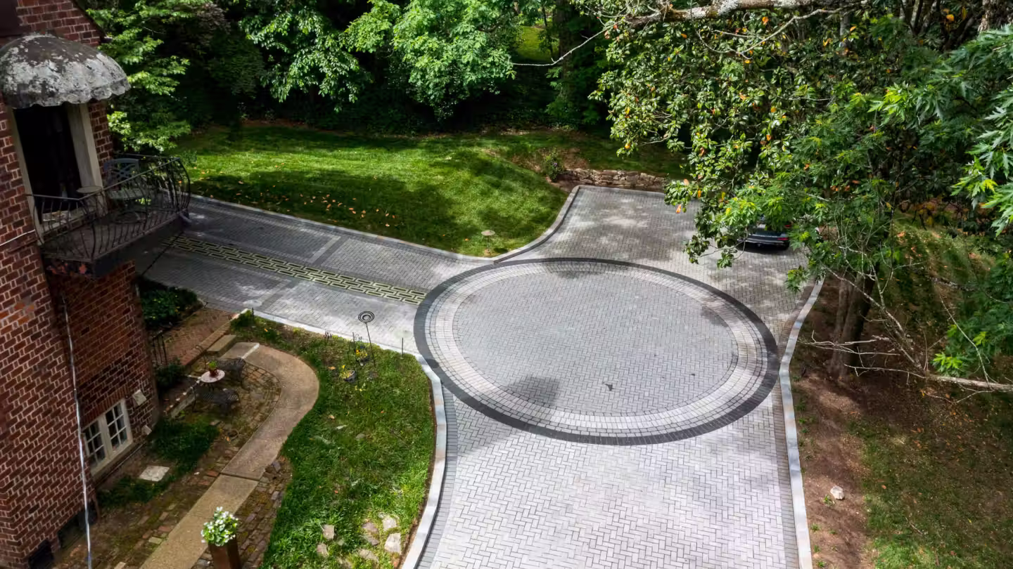 Aerial view of a circular patterned driveway with surrounding green trees, lawn, and a brick building with a balcony.