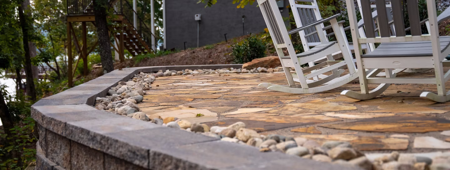 Stone patio with white wooden rocking chairs and decorative rocks along raised stone wall in a wooded backyard.