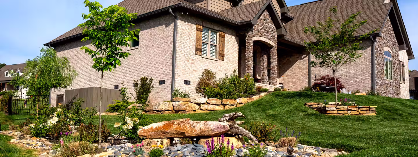 Modern brick house with a manicured lawn, young trees, and a landscaped rock garden with flowers.