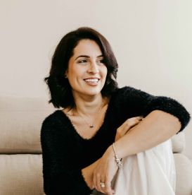A dark haired woman smiling sitting on a sofa holding her knee