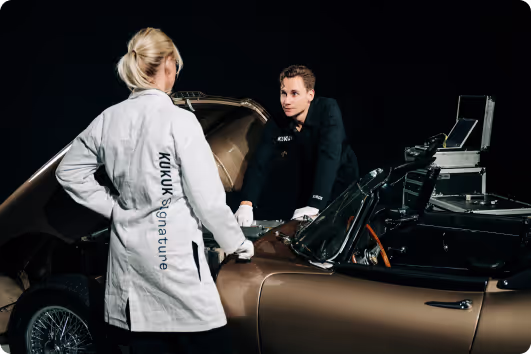 A woman in a white lab coat labeled 'KUKU Signature' and a man in black gloves inspecting the interior of a classic convertible car against a dark background.