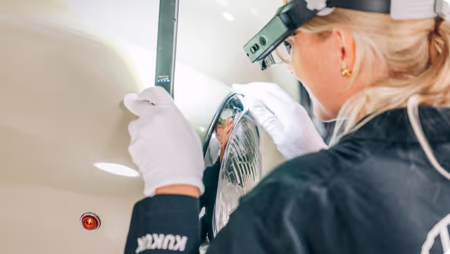 Technician wearing white gloves inspecting a vehicle headlight closely with a magnifying tool.